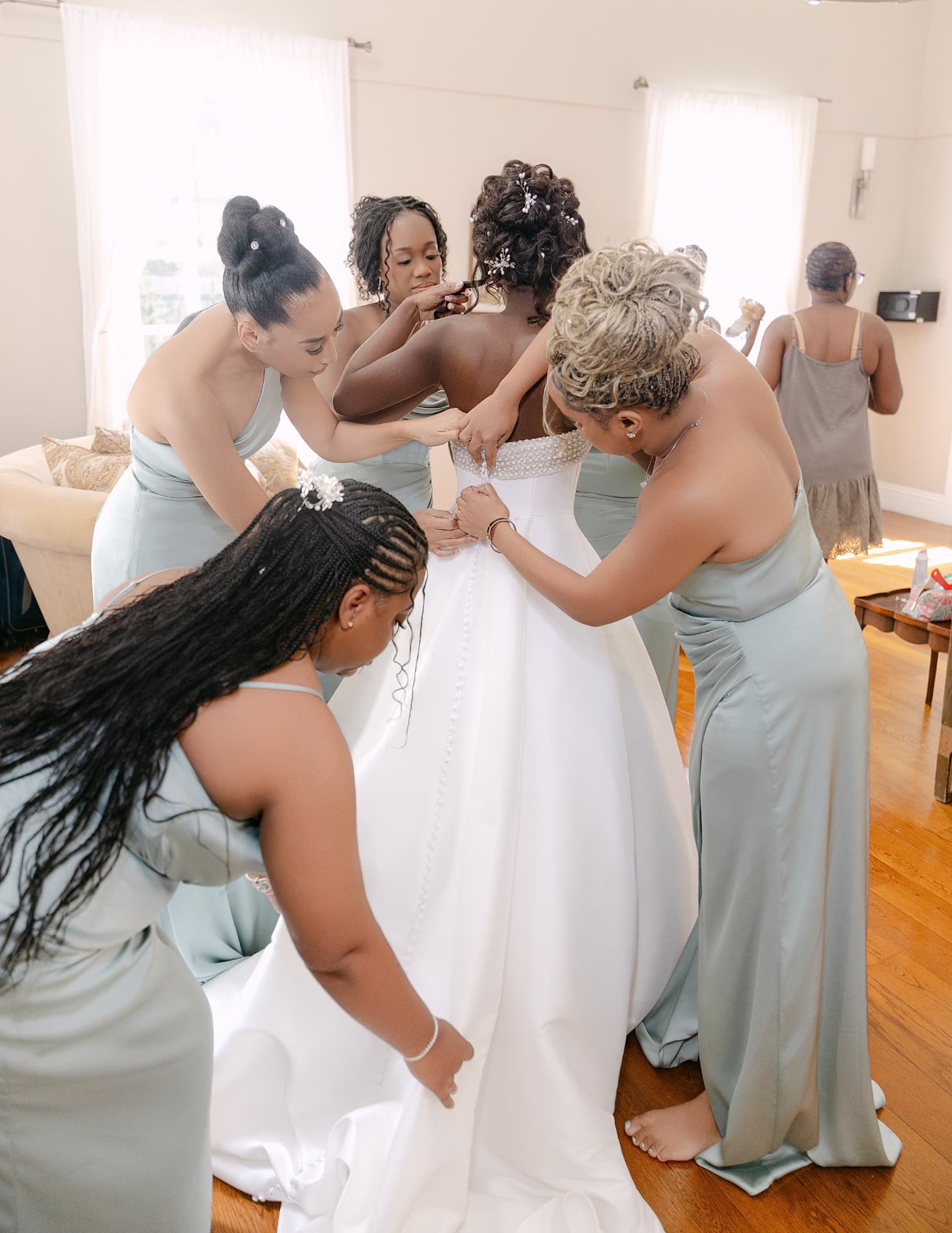 Bridesmaids in sage gowns fastening the bride's buttons in a sunlit suite