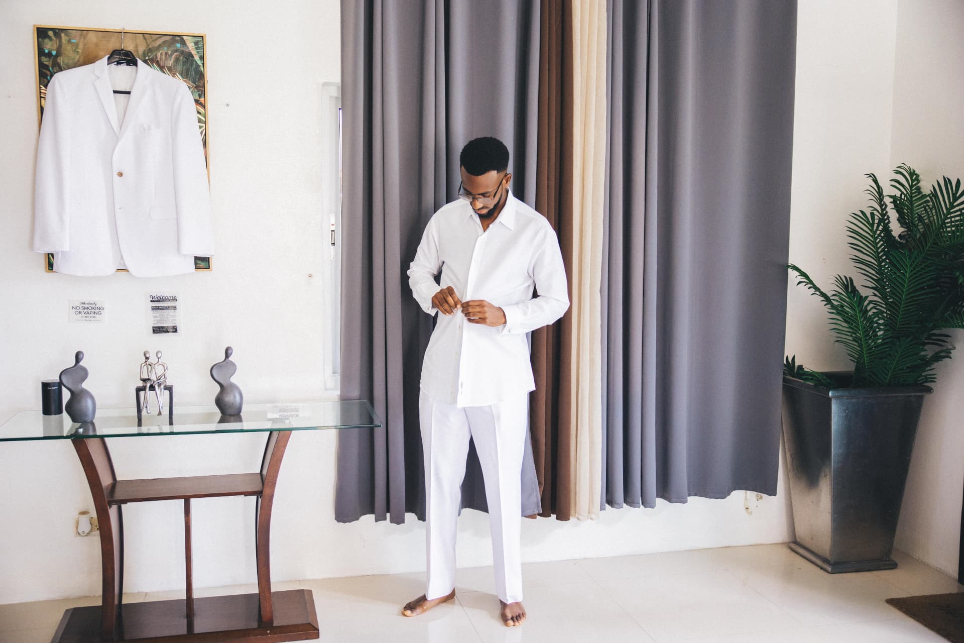 Groom buttoning his white shirt with his jacket hanging on the wall