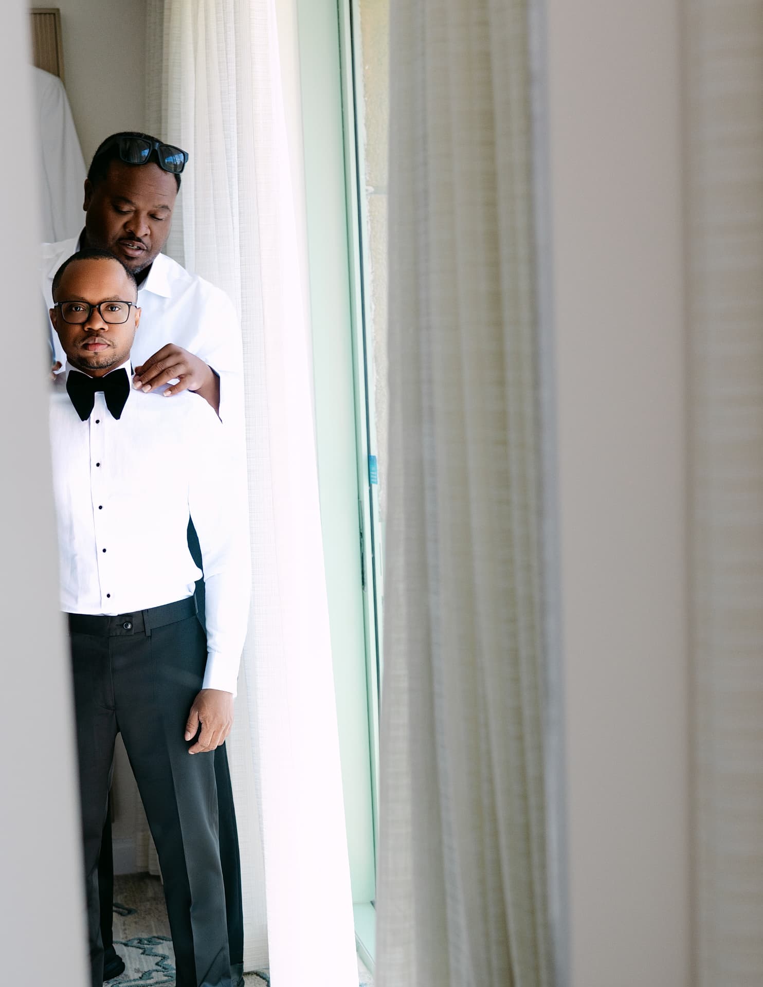 Groom standing by sheer curtains while a groomsman checks his collar