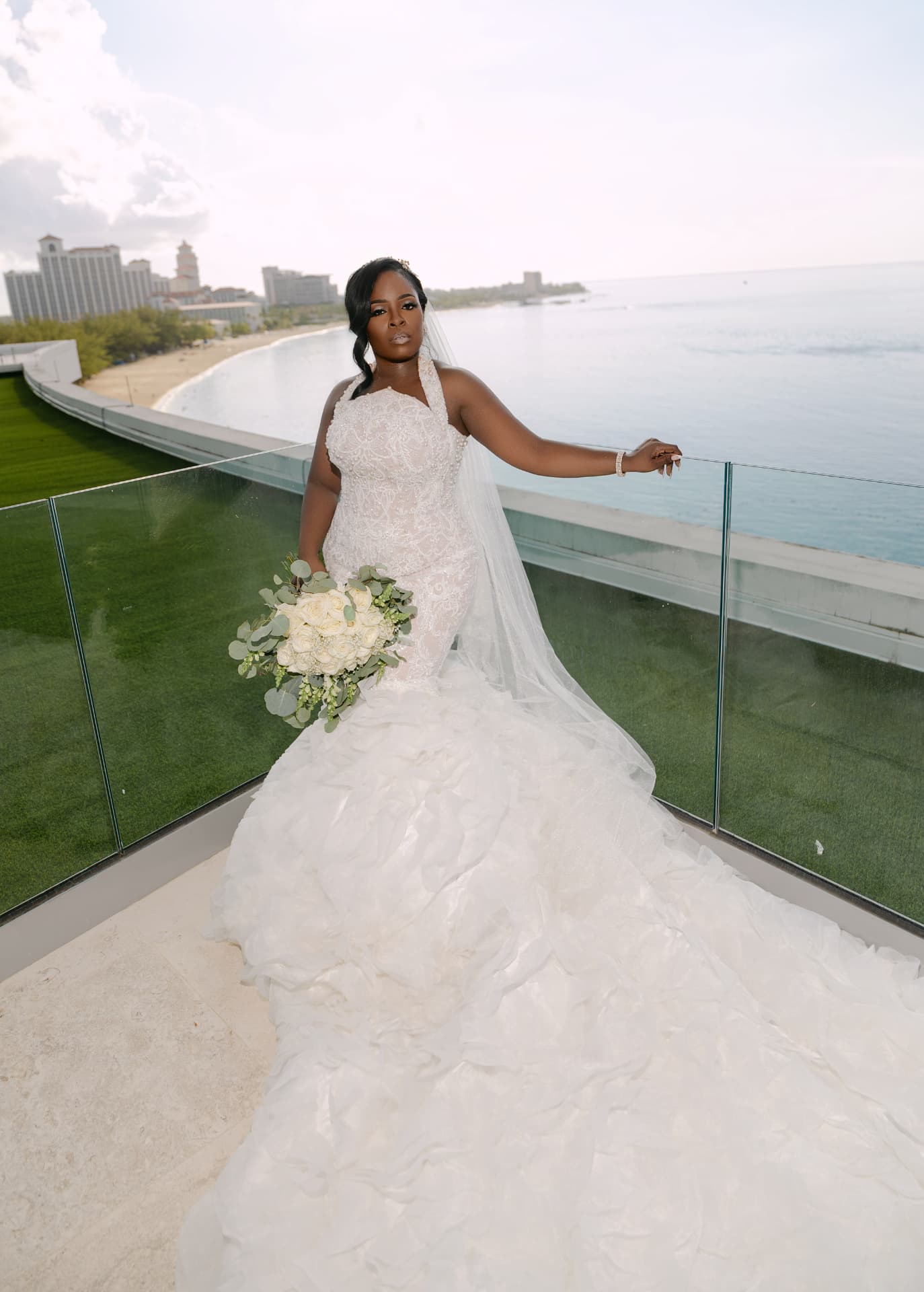 Bride in her lace gown with bouquet and veil, standing on a glass terrace overlooking the ocean