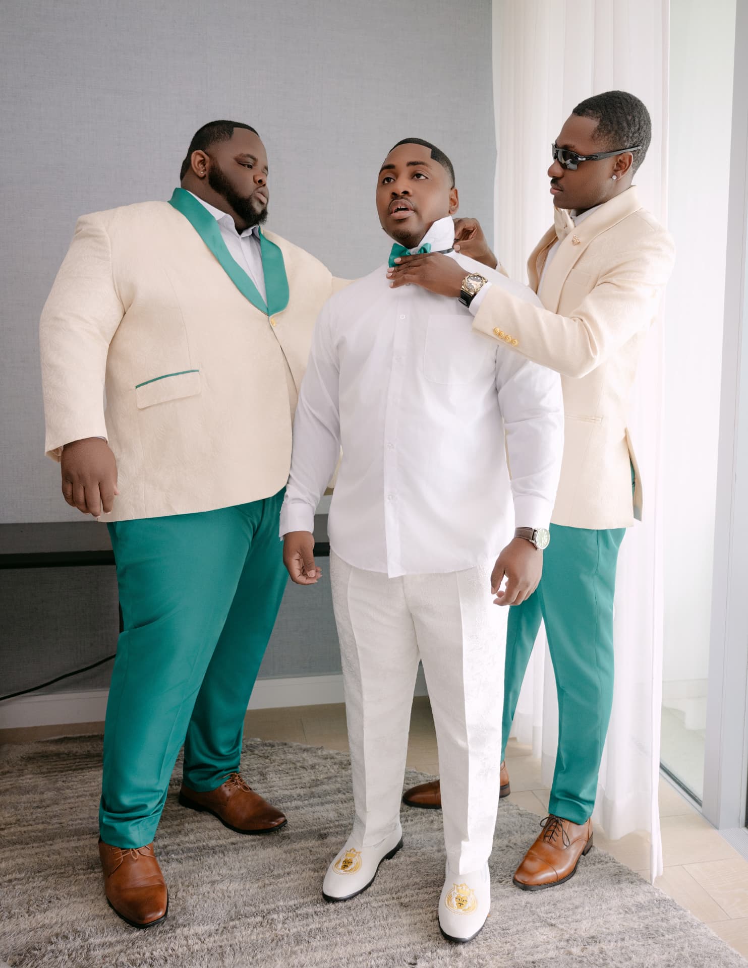 Two groomsmen in cream and teal suits tying the groom's bow tie