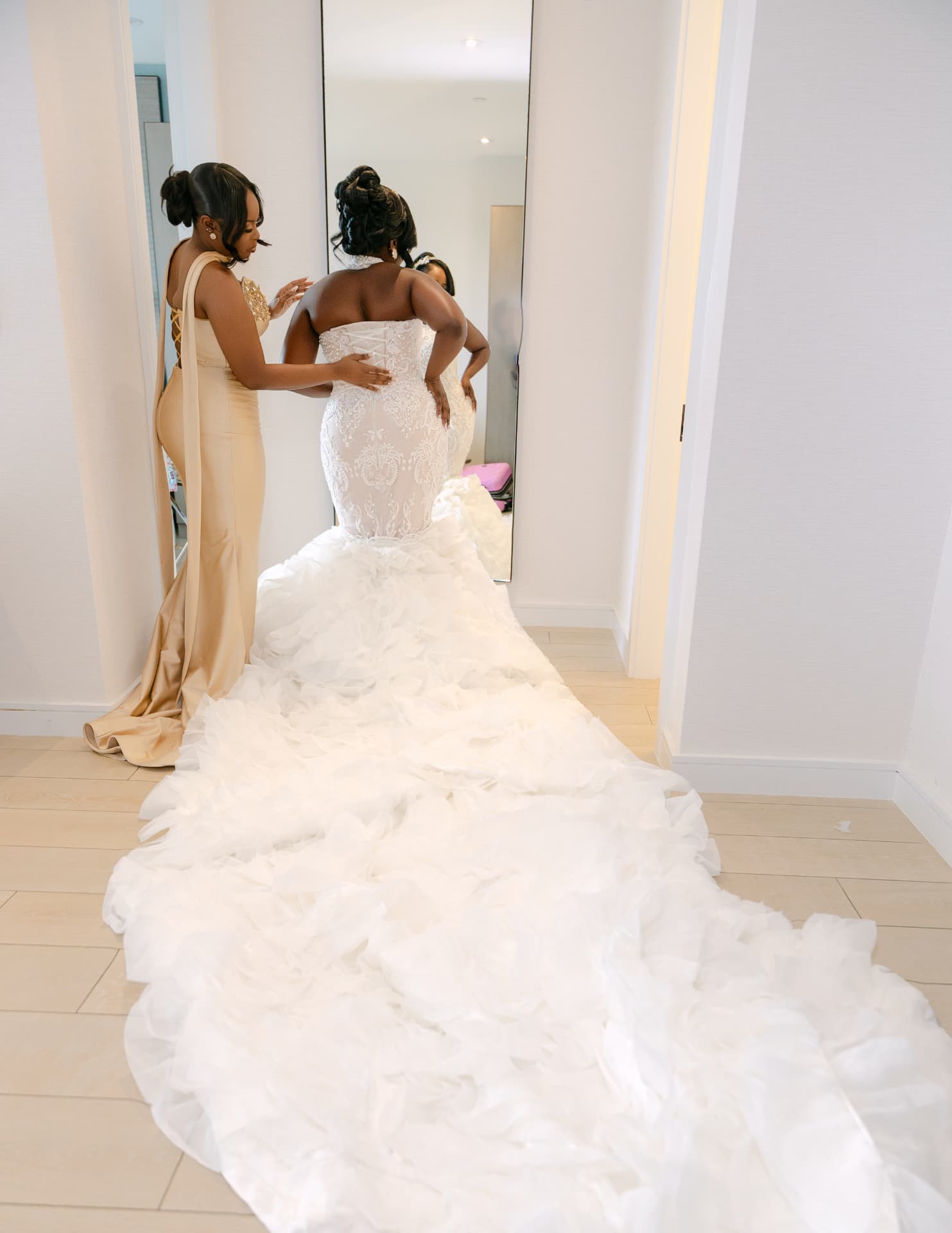 Bridesmaid fastening the bride's gown in front of a mirror, long ruffled train trailing
