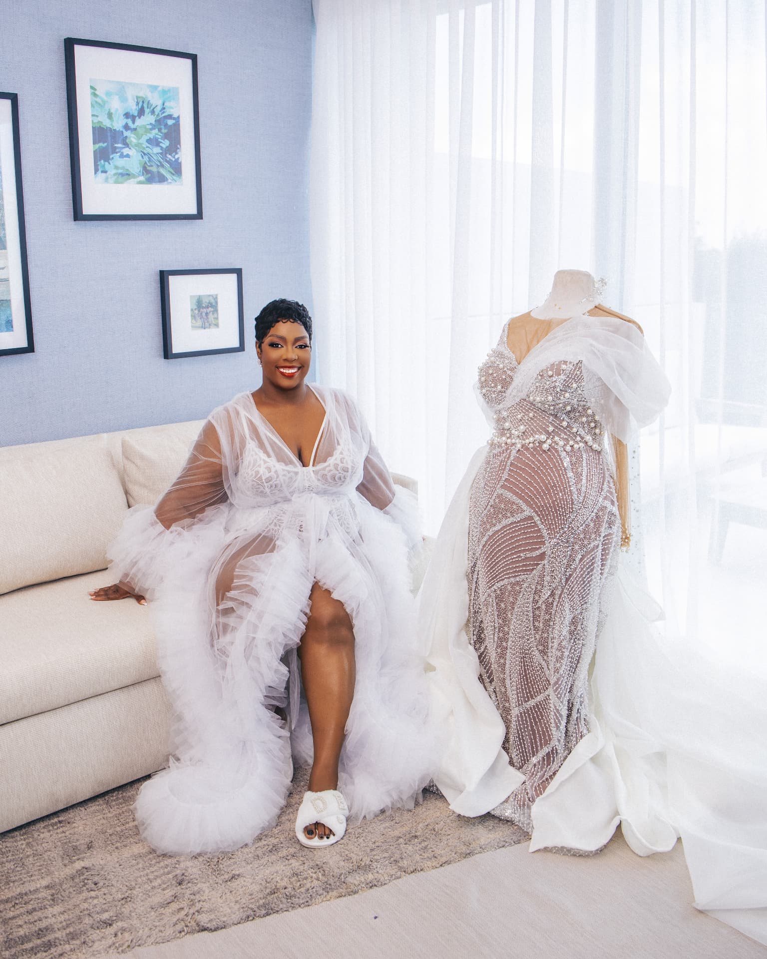 Bride seated in a tulle robe beside her beaded gown on a dress form